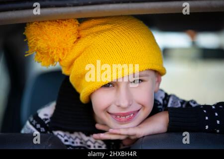 Positive little boy in a yellow knitted hat looks at the camera and smiles. Portrait of a ten year old boy. Child in the car window. Stock Photo