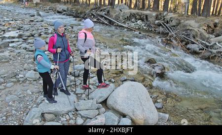 Woman do Nordic walking in nature near mountain river. Girls and children use trekking sticks and nordic poles, backpacks. Family travels sports. Kid Stock Photo