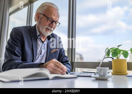 Mature businessman or entrepreneur working on laptop computer and taking notes and writing. Handsome senior business leader working in a modern corpor Stock Photo