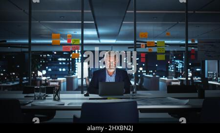 Successful Senior Businessman Working on Laptop Computer in Big City Office Late in the Evening. Investment Analyst Checking Financial Graphs from Project Management Reports. Stock Photo