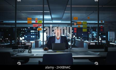 Successful Senior Businessman Working on Laptop Computer in Big City Office Late in the Evening. Investment Analyst Checking Financial Graphs from Project Management Reports. Stock Photo