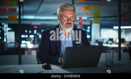 Successful Senior Businessman Working on Laptop Computer in Big City Office Late in the Evening. Investment Analyst Checking Financial Graphs from Project Management Reports. Stock Photo