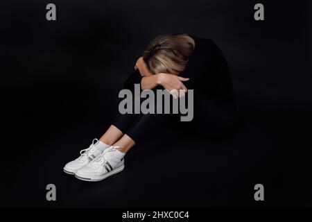 Young blonde woman in black dress going through depression with face on knees sitting on floor hiding from people on black background. Physical and Stock Photo