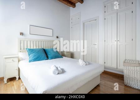 King size bed with white wooden headboard, wall covered with white cabinet doors, blue cushions and wicker laundry basket Stock Photo