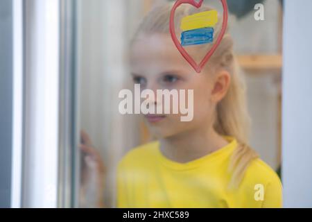 Girl looking out the window, upset and survives the war Ukraine Russia, flag of Ukraine is painted on the window Stock Photo