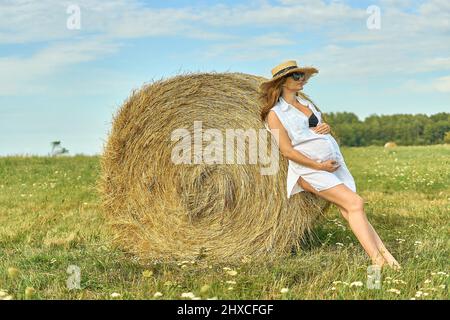 A beautiful young pregnant woman in the hat stands on a green field, leaning against a haystack. Sunny summer day. She is happy. Stock Photo