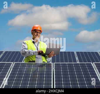 Mature male engineer using a laptop computer on a roof with solar panels and smiling at camera Stock Photo