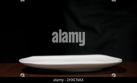 White plate of square shape. The dishes are on a black background on a brown table. Side view. Stock Photo