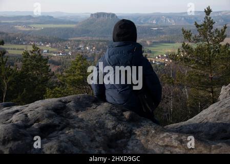 A boy sits on the Papststein and looks over to the Lilienstein, table mountain in the Elbsandsteingebirge, located at the Malerweg, Papstdorf, Saxony, Germany Stock Photo