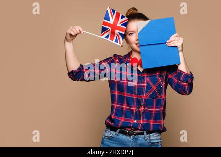 Funny woman peeks out from behind open book, half face visible. Student girl holds small UK flag on beige background. Study abroad. International student exchange program. Learning British English. Stock Photo