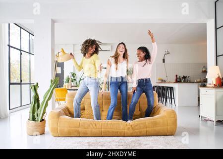 Three rebellious women friends dancing and singing on the sofa. Cheerful girls enjoy together at home having fun.  Stock Photo