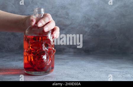 flacon with red liquid in a woman's hand on a dark background. bottle in the shape of a human skull.  Stock Photo