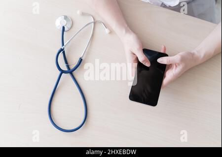 Unrecognizable female doctor sits at a desk and writes a message on a smartphone. Practitioner holds a phone in his hands and answers patients Stock Photo