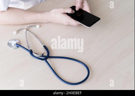 Unrecognizable female doctor sits at a desk and writes a message on a smartphone. Practitioner holds a phone in his hands and answers patients Stock Photo