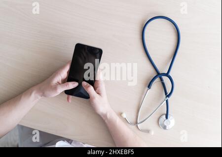 Unrecognizable female doctor sits at a desk and writes a message on a smartphone. Practitioner holds a phone in his hands and answers patients Stock Photo