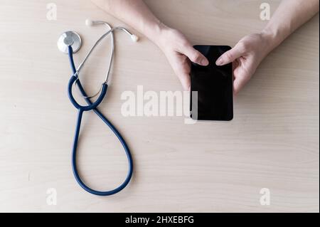 Unrecognizable female doctor sits at a desk and writes a message on a smartphone. Practitioner holds a phone in his hands and answers patients Stock Photo