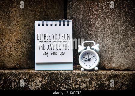 Motivational quote text on notepad - Either you run the day or the day runs you. Rock wall background. Motivational concept Stock Photo