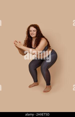 Vertical image of glad ginger massive big obesity fat overweight woman, in gray leggings and top. Knee bending, squat Stock Photo