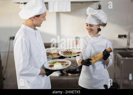 Chef standing with ready meals, talking with female cook in the kitchen. Happy caucasian cook working at restaurant. Concept of professional occupation and teamwork Stock Photo