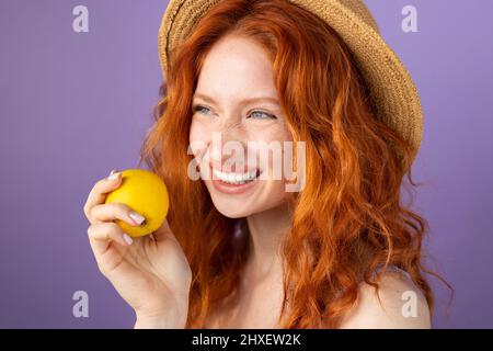 Happy smiling redhead pretty young woman wearing straw hat holding an apple in her hand to maintaining helthy lifestyle. Stock Photo