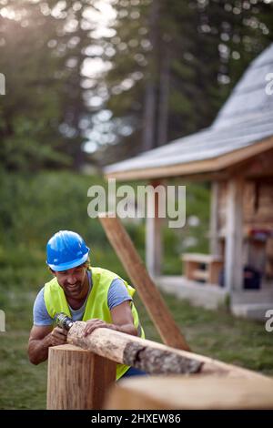 Young worker working with drilling machine on wooden construction. Carpenter building wooden home Stock Photo