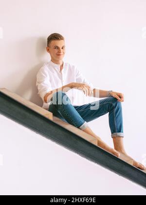 handsome guy in white shirt and blue jeans sitting on a stairs indoor. Fashion, style concept Stock Photo