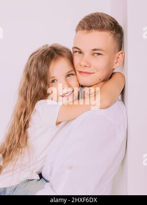 caucasian happy adorable siblings - teenager boy brother and little girl sister cuddling on white background indoor. Family, relationships, relatives, Stock Photo