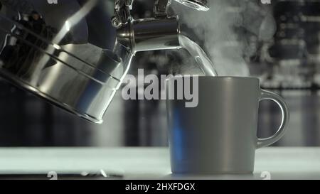 Close up of making hot beverage tea or coffee at the kitchen at home. Action. Pouring hot water from the metal teapot inside matte ceramic mug. Stock Photo