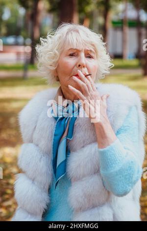sad fashionable senior woman with grey hair in fur coat outdoor smoking cigarette. Unhealthy lifestyle, age, oldness, addiction, bad habit concept. Stock Photo