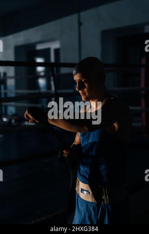 Vertical shot of confident boxer wrapping defense boxing tape around arms before fight in sport club with dark interior on background of ring Stock Photo