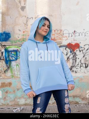 Teenager wearing light blue oversize hoodie and jeans looking into the camera. Blue haired teen girl stays outdoors against graffiti wall. Clothing mo Stock Photo