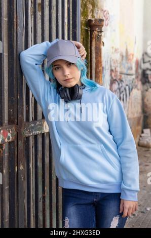 Teenager wearing light blue oversize hoodie and baseball cap with hand over head. Blue haired teen girl with wireless headphones stays outdoor near a Stock Photo