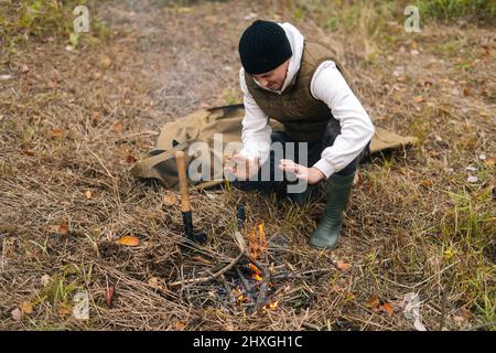 High-angle view of frozen tourist male wearing warm clothes warming hands over fire at outdoors on overcast cold day. Stock Photo