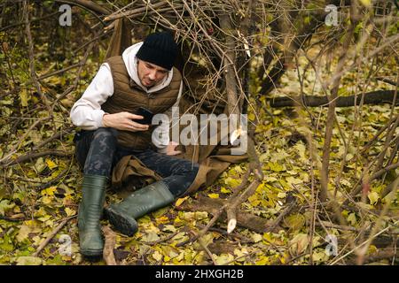 Tired traveler male wearing warm clothes sitting in forest and using typing mobile phone in overcast cold day. Stock Photo