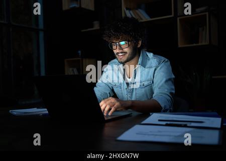 Smiling young indian business man working online late at night using laptop. Stock Photo