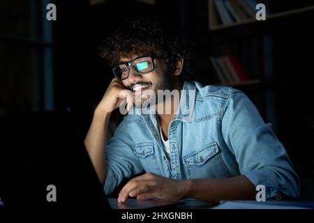 Happy young indian man working online late at night using laptop computer. Stock Photo