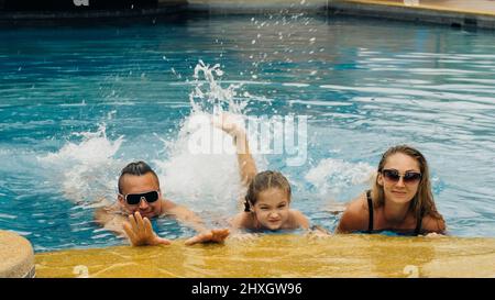The mother and father with little daughter have fun in the pool. Mom and dad plays with the child. The family enjoy summer vacation in a swimming pool