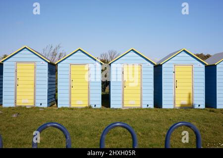 Yellow and blue beach huts seen behind a green lawn and an iron railing in Felpham, West Sussex, UK. Stock Photo