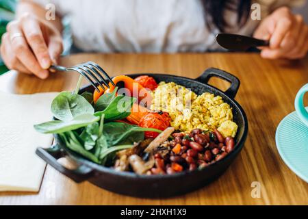 Vegan dish from different vegetables on frying pan and woman hands with fork and knife at table in cafe closeup Stock Photo