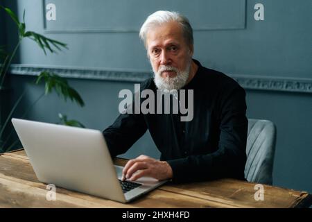 Medium shot of handsome gray-haired mature business man sitting at table with laptop computer at home office, looking at camera. Stock Photo