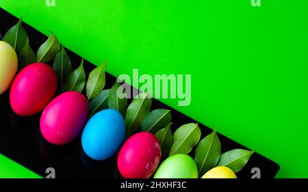 Easter. Brightly colored eggs and green young leaves on a black rectangle. Corner for text. Green background. Stock Photo