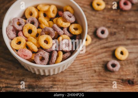 Colorful round fruit flakes in a white bowl scattered on a brown wooden table, top view Stock Photo