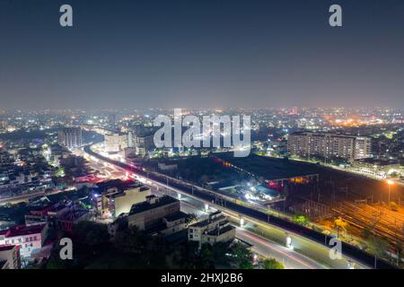 elevated metro track going across the screen over a lit road surrounded by buildings and metro train yard in distance with cityscape lights Stock Photo