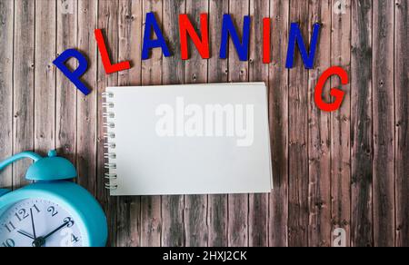 The word planning in wooden colored letters over a notepad with a clock on a wooden table. Stock Photo