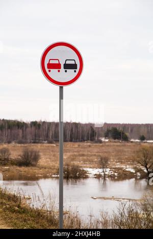 The overtaking sign is prohibited on the background of the river and meadow. Spring, the snow is melting, there are puddles of slush and mud all around. Day, cloudy weather, soft warm light. Stock Photo