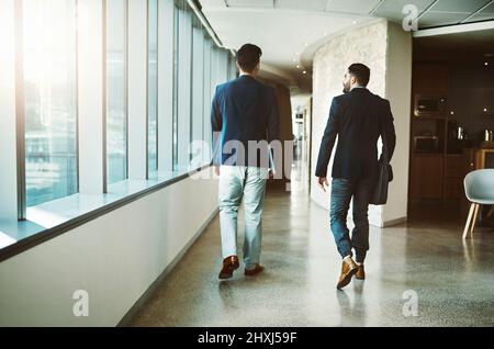 Success doesnt happen until people get moving. Rearview shot of two young businessmen walking down a corridor of a modern office. Stock Photo