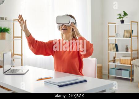 Amazed woman in virtual reality headset touches air while sitting at her working place in office. Stock Photo