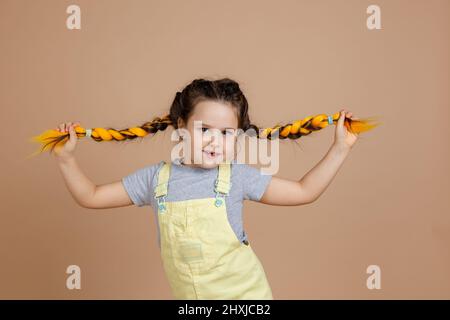 Portrait of indulding overjoyed small girl holding kanekalon braids with hands, smiling with missing tooth looking at camera wearing yellow jumpsuit Stock Photo