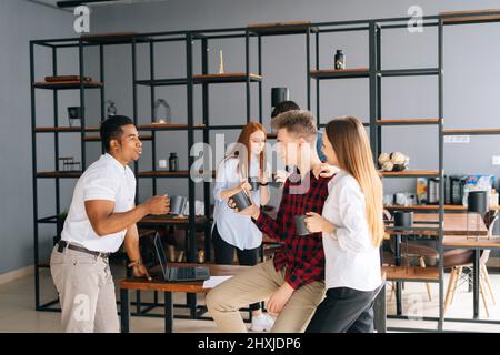 Group of five happy young business multiethnic start-up colleagues enjoying pleasant conversation during coffee break in co-working space. Stock Photo