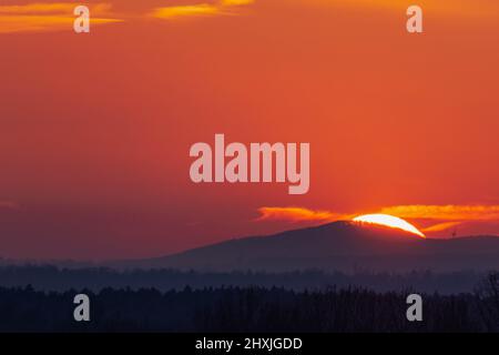 The top of the mountain is hidden in the twilight. Behind the mountain you can see. partly behind it, a red shield of the setting sun is hidden. Stock Photo
